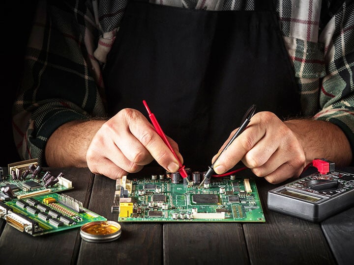 Electrician in a plaid shirt and black apron testing a circuit board assembly with an electric multi-tester tool