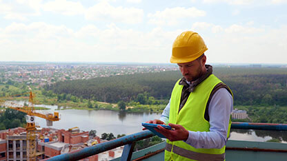 Focused Caucasian young man in uniform and helmet working on digital tablet while standing on construction site. Competent engineer checking working process. Building industry.