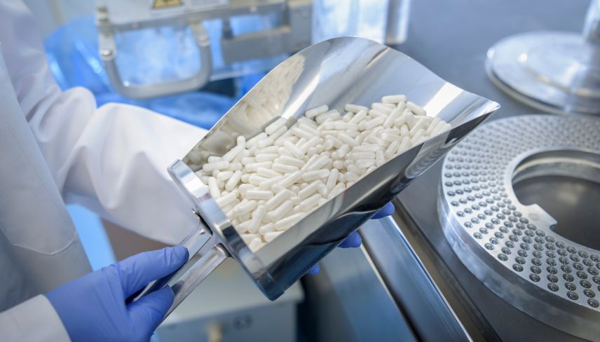 Gloved technician holding a scoop of white capsules in a pharmaceutical manufacturing facility.